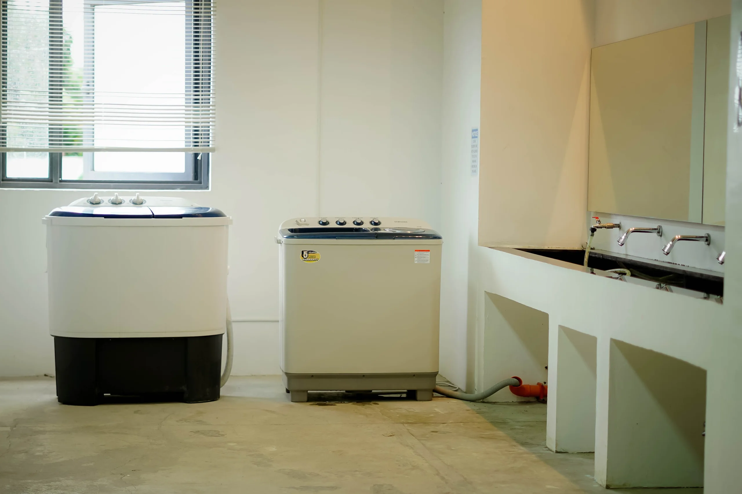 窓のある部屋に並ぶ2台の二層式洗濯機と、壁ぞいの洗い場